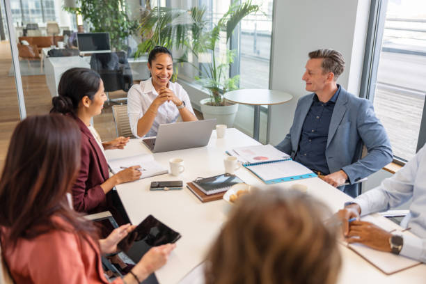 Aerial shot of a pleasant and interesting meeting where there are workers of different ethnic origins, where some use their electronic devices or notebooks to take notes while having cups of coffee in a bright day in the conference room.